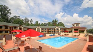 Zona de piscina de hotel con sillones de descanso y sombrillas, rodeada por un edificio con varias ventanas y balcones.
