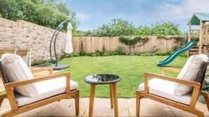 Un patio trasero con una estructura de juego de madera, un tobogán y dos sillas acolchadas con una pequeña mesa.