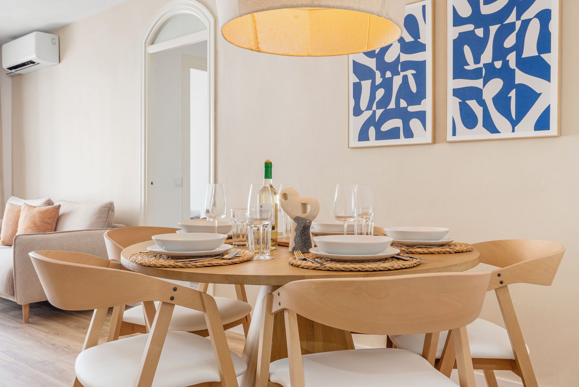 A dining area with a round table set for four, surrounded by wooden chairs with white cushions. There are plates, glasses, and a bottle of wine on the table. A woven placemat is under each plate. The room has a neutral color scheme with a beige sofa in the background.