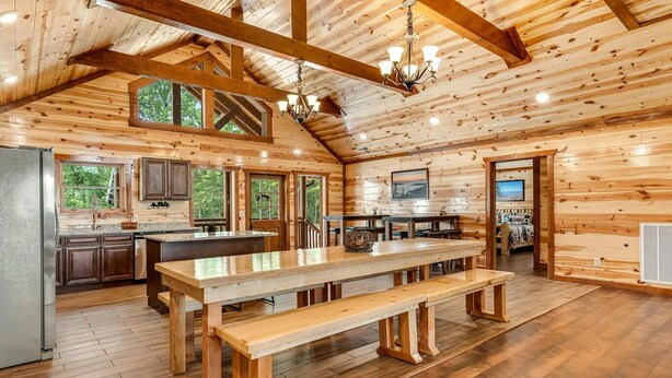 Une vaste cabane en bois dotée d’une grande table à manger, d’un coin cuisine et de nombreuses fenêtres offrant une vue sur la forêt environnante.