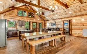 Une vaste cabane en bois dotée d’une grande table à manger, d’un coin cuisine et de nombreuses fenêtres offrant une vue sur la forêt environnante.