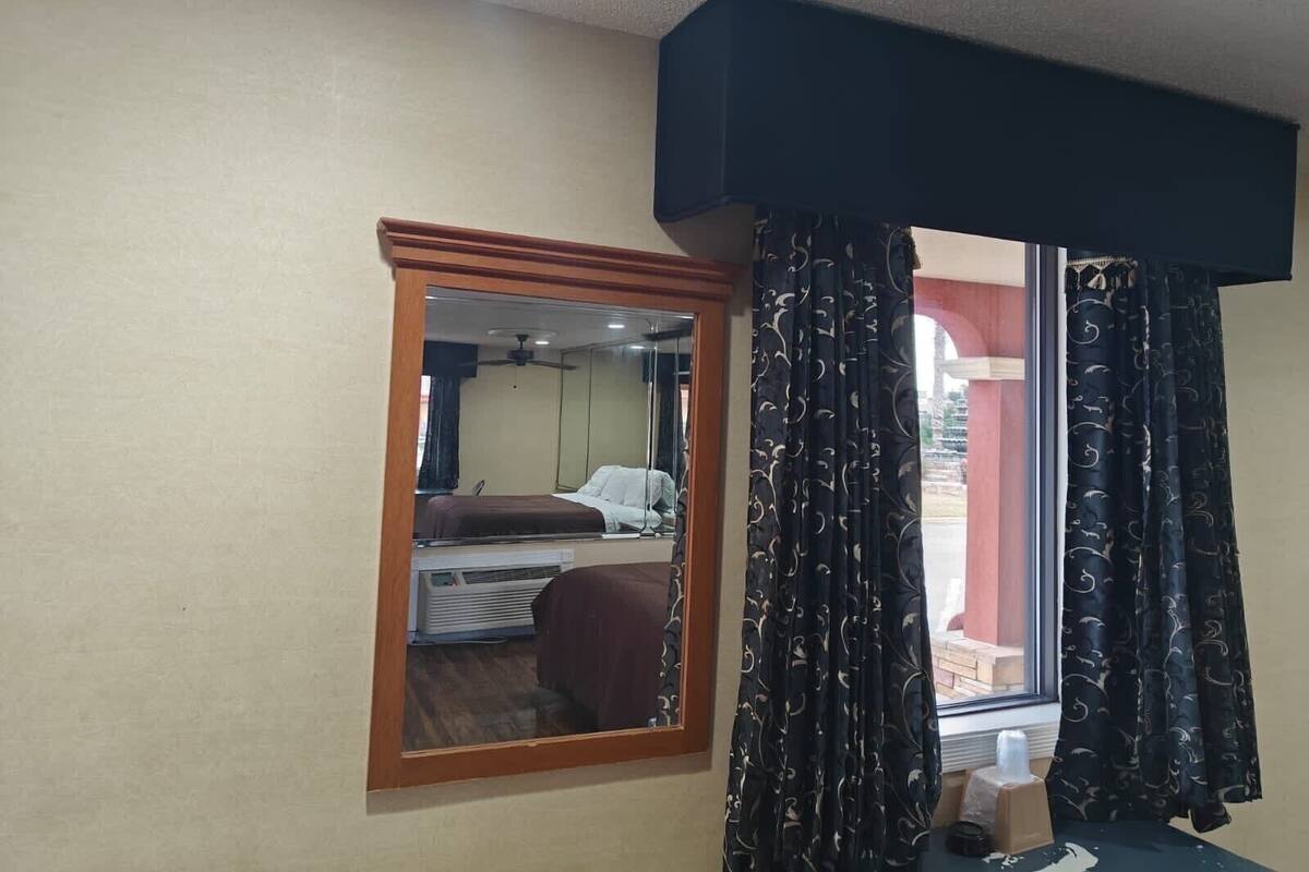 A hotel room with a bed, a window with curtains, a mirror, and a ceiling fan.