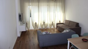 Una sala de estar moderna con un sofá gris, una mesa de comedor de madera y un televisor sobre un mueble blanco.