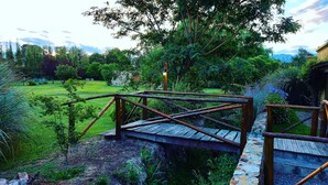 A wooden bridge over a stone path leading to a wooden deck, surrounded by lush greenery and a clear sky. - Posada del Santo Green & Rest (Cerrillos)