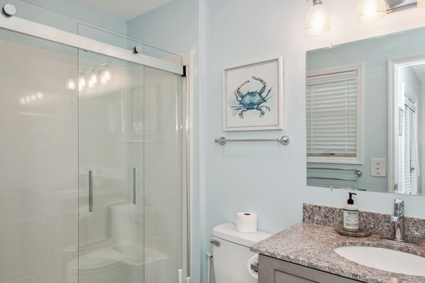 A bathroom with a glass shower enclosure, a toilet, a sink with a marble countertop, a framed artwork of a crab, and a hand soap dispenser.