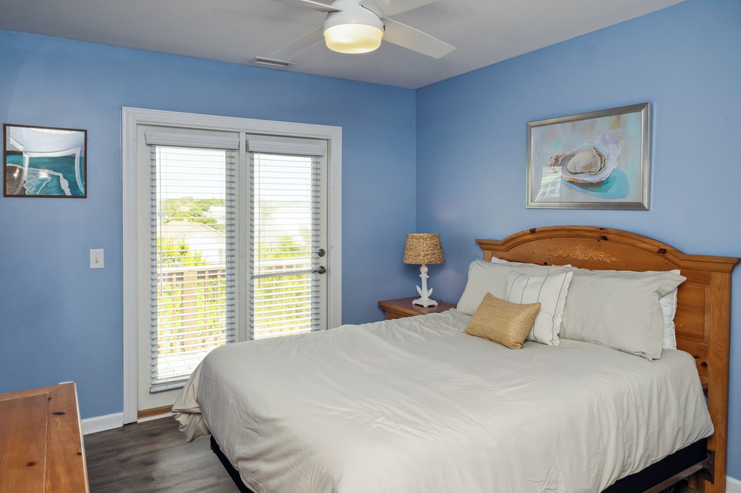 A bedroom with a bed, a nightstand, a ceiling fan, a window with blinds, and two framed pictures on the wall.