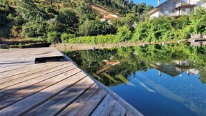 A wooden deck leading to a calm water body with reflections, surrounded by lush greenery and residential buildings. - Casinhas do Amioso T1 in the village of Amioso do Senhor in Alvares - Góis (Góis)