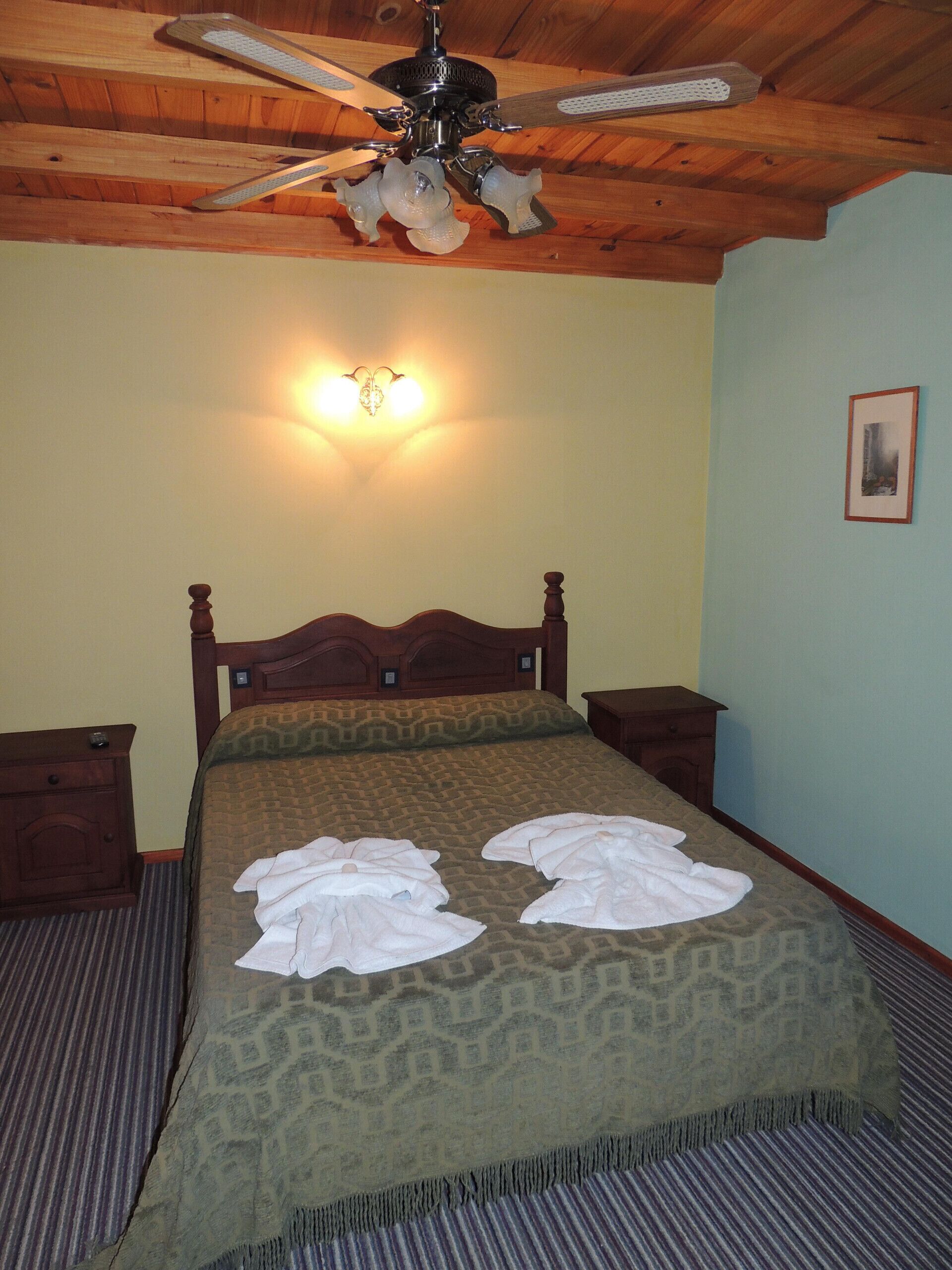 A hotel room with a wooden ceiling, a bed with a patterned bedspread, two bedside tables, and a ceiling fan.