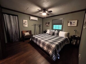 A bedroom with a bed, a ceiling fan, an air conditioning unit, a window with blinds, and a nightstand with a lamp. - Feisty Ranch Farm Retreat - Private and charming 2-bedroom on 15 acres (Aquone)