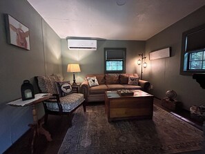 A cozy living room with a sofa, armchair, and coffee table, a lamp, and a window with a view. - Feisty Ranch Farm Retreat - Private and charming 2-bedroom on 15 acres (Aquone)