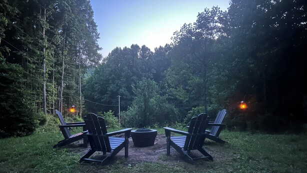Un área de estar al aire libre con sillas Adirondack y una fogata rodeada de árboles.