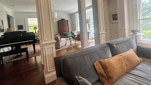Un amplio salón con un piano de cola, un sofá gris con un cojín marrón, y vistas a un jardín exuberante a través de grandes ventanales.