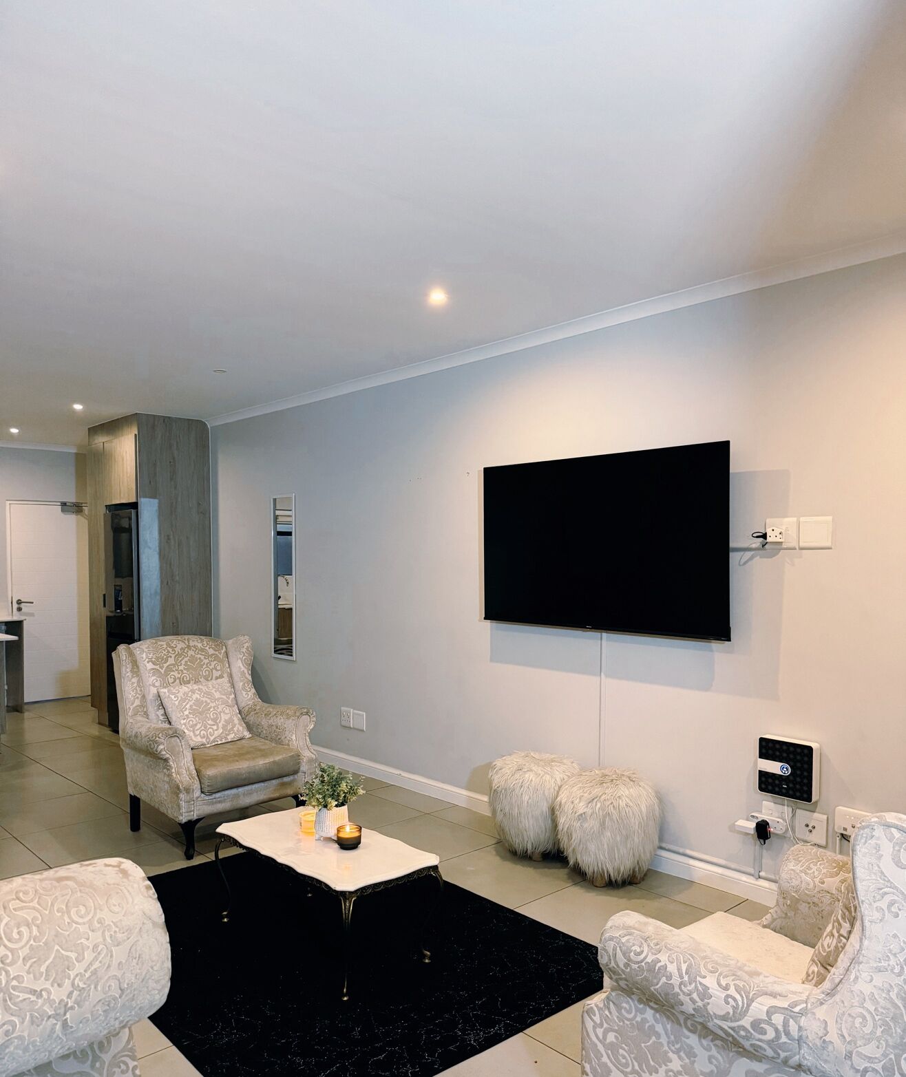 Ein modernes Wohnzimmer mit einem an der Wand montierten Flachbildfernseher, einem gemusterten Sessel, einem Couchtisch mit Dekoration und einem kuscheligen Ottomanen.