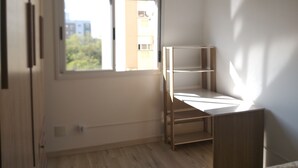 Una habitación pequeña y luminosa con una ventana, un escritorio de madera y una pared blanca.