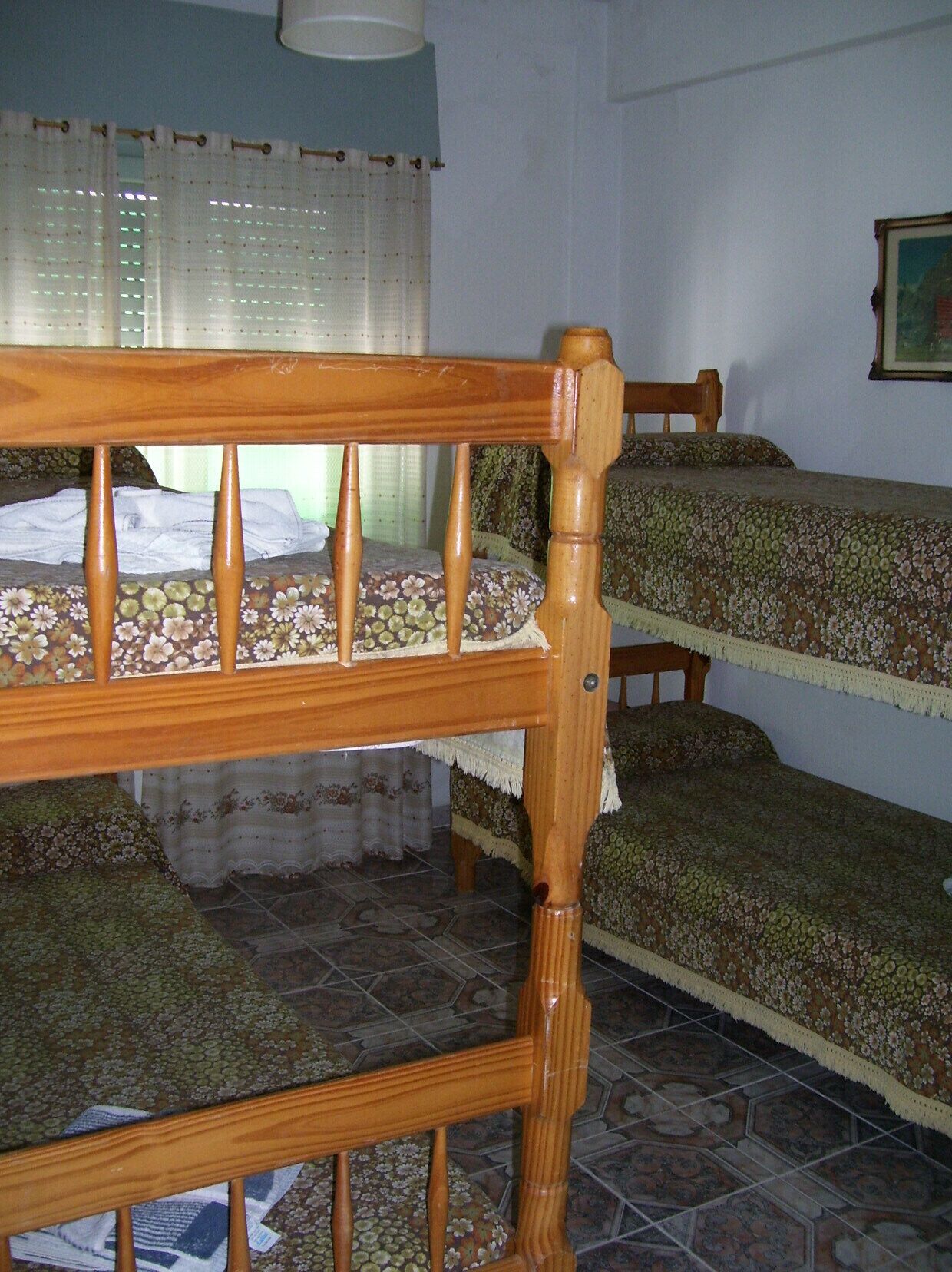 Estructura de cama de madera con una colcha estampada con flores, una ventana con cortinas y un piso de baldosas.