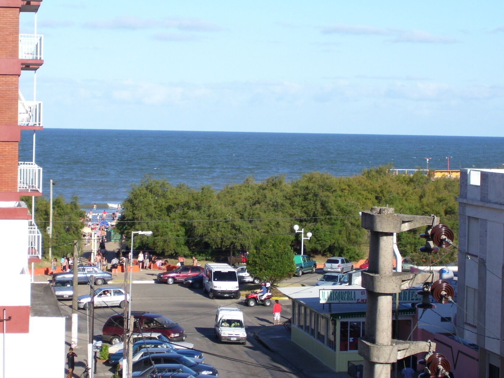 A coastal street with parked cars, a visible building with a sign, and a view of the sea.