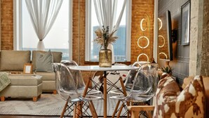 A modern living room with a sofa, clear chairs, a dining table, and a vase with dried plants. - Rodeo Room Downtown Loft | Skyline View, Pool ,Gym & Free Valet Parking (Dallas)