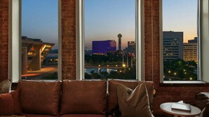 A room with a brick wall, a brown sofa, and a view of a cityscape through large windows. - Rodeo Room Downtown Loft | Skyline View, Pool ,Gym & Free Valet Parking (Dallas)