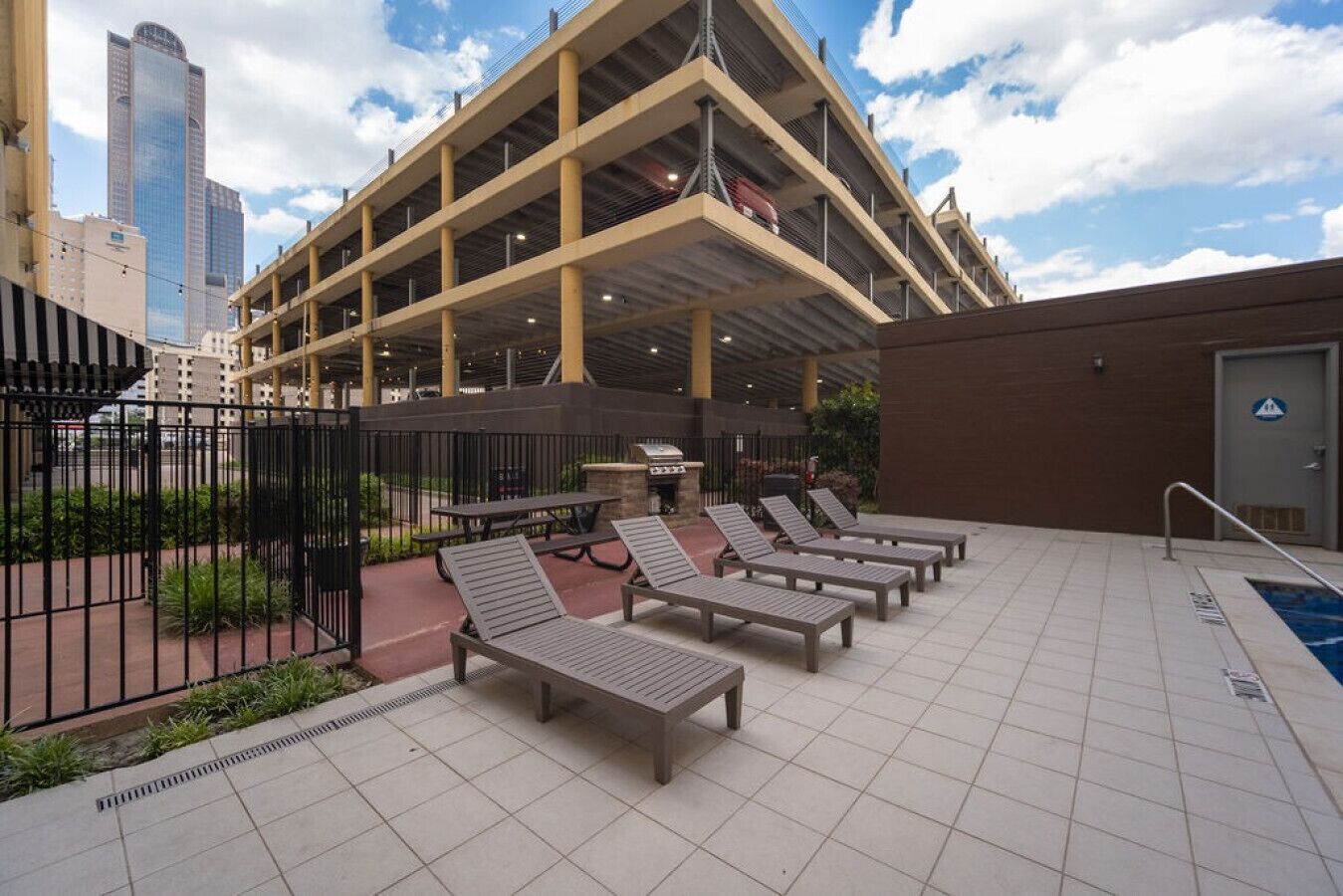 A rooftop pool area with lounge chairs, a barbecue grill, and a view of a parking garage.