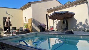 Une piscine avec un espace de détente comprenant des chaises et un parasol, ainsi qu’un bâtiment doté d ’un toit en tuiles.