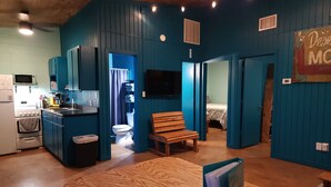 A compact room with a kitchenette, a wooden table, a blue armchair, and a bed in the adjacent room.