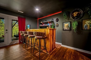 Uma área de bar com banquetas, balcão de madeira e prateleiras com copos e garrafas.