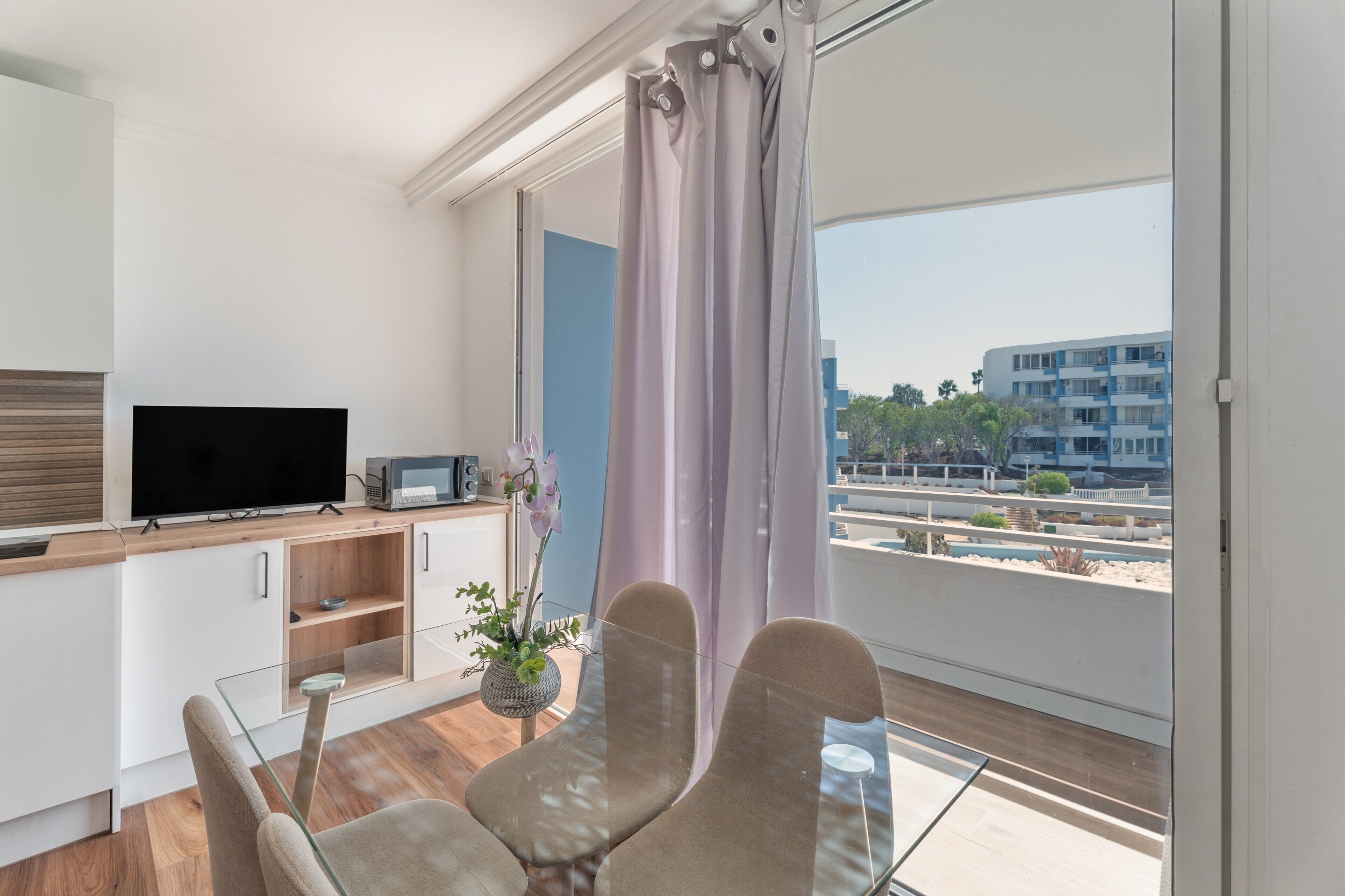 A modern living room with a flat-screen TV, a glass table, two chairs, a small cabinet, and a view of a building through a window with curtains.