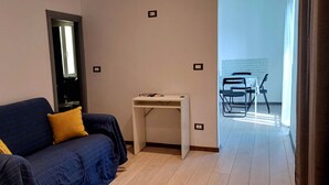A room with a dark blue sofa, a small white table, and a view of a dining area with chairs and a table. - Al Porto Antico (Genoa)