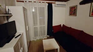 Un salón con televisión, una mesa blanca, un sofá rojo y una ventana con cortinas translúcidas.