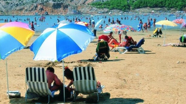 Una playa con gente tomando el sol y nadando, sombrillas y una ladera rocosa al fondo.