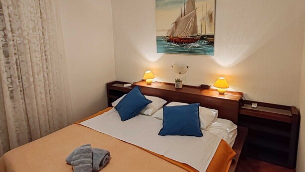 A neatly made bed with a beige bedspread and two blue pillows, a painting of a sailboat on the wall, and two bedside lamps on wooden nightstands.