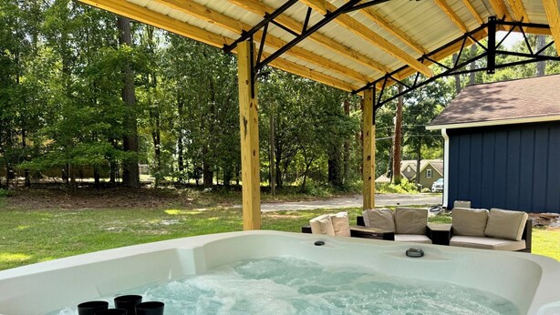 A hot tub under a wooden pergola with a view of a backyard and a house.