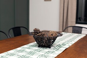 A dining table with a woven centerpiece containing pine cones, set on a table runner with a leaf pattern.