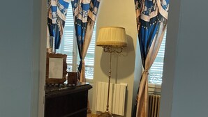 Une pièce avec un plancher en bois, un radiateur, des rideaux, une lampe et un meuble.