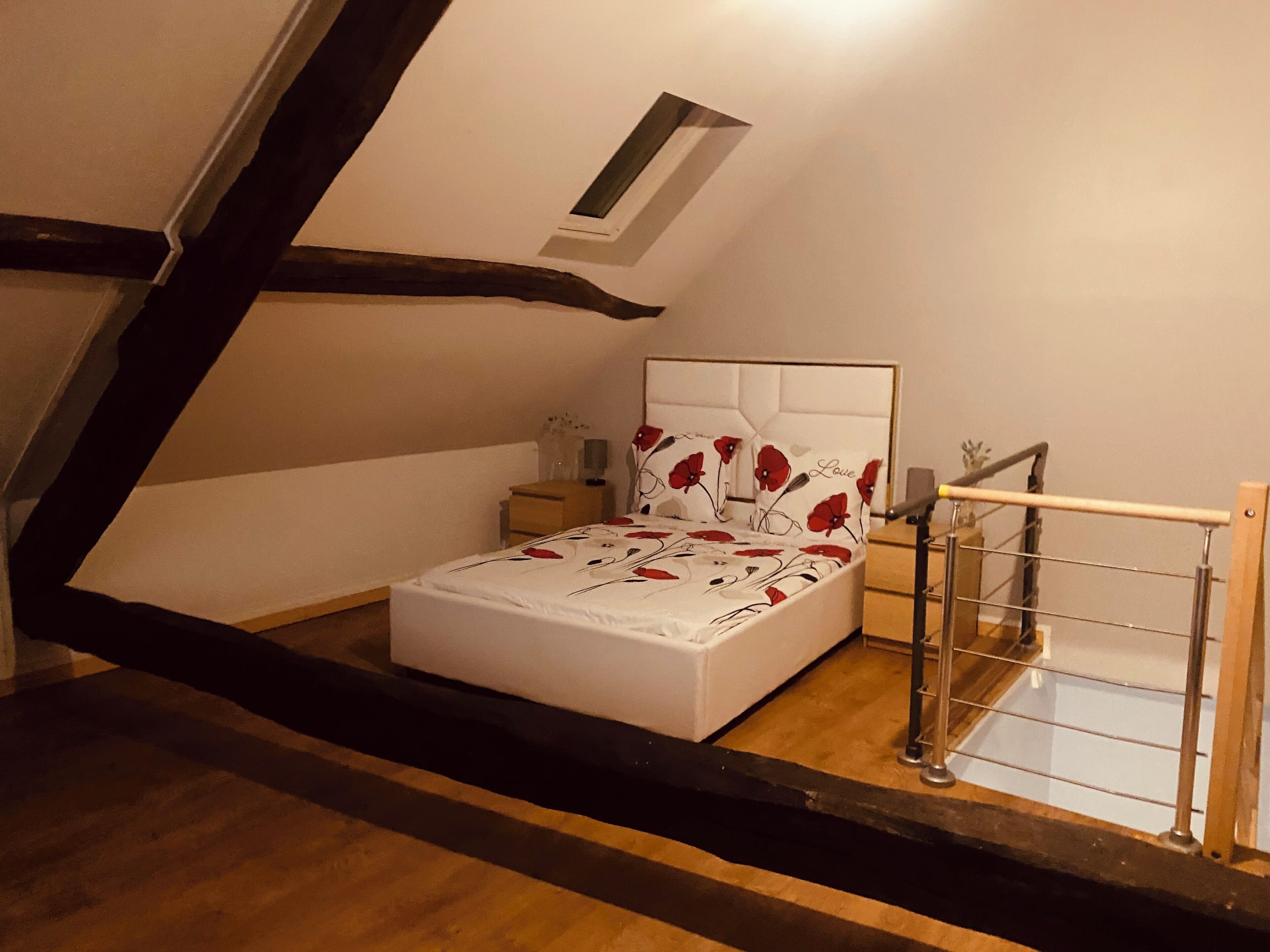 Een slaapkamer met een schuine kap, een bed met bloemenbedekking, een nachtkastje, een klein tafeltje en een metalen leuning.