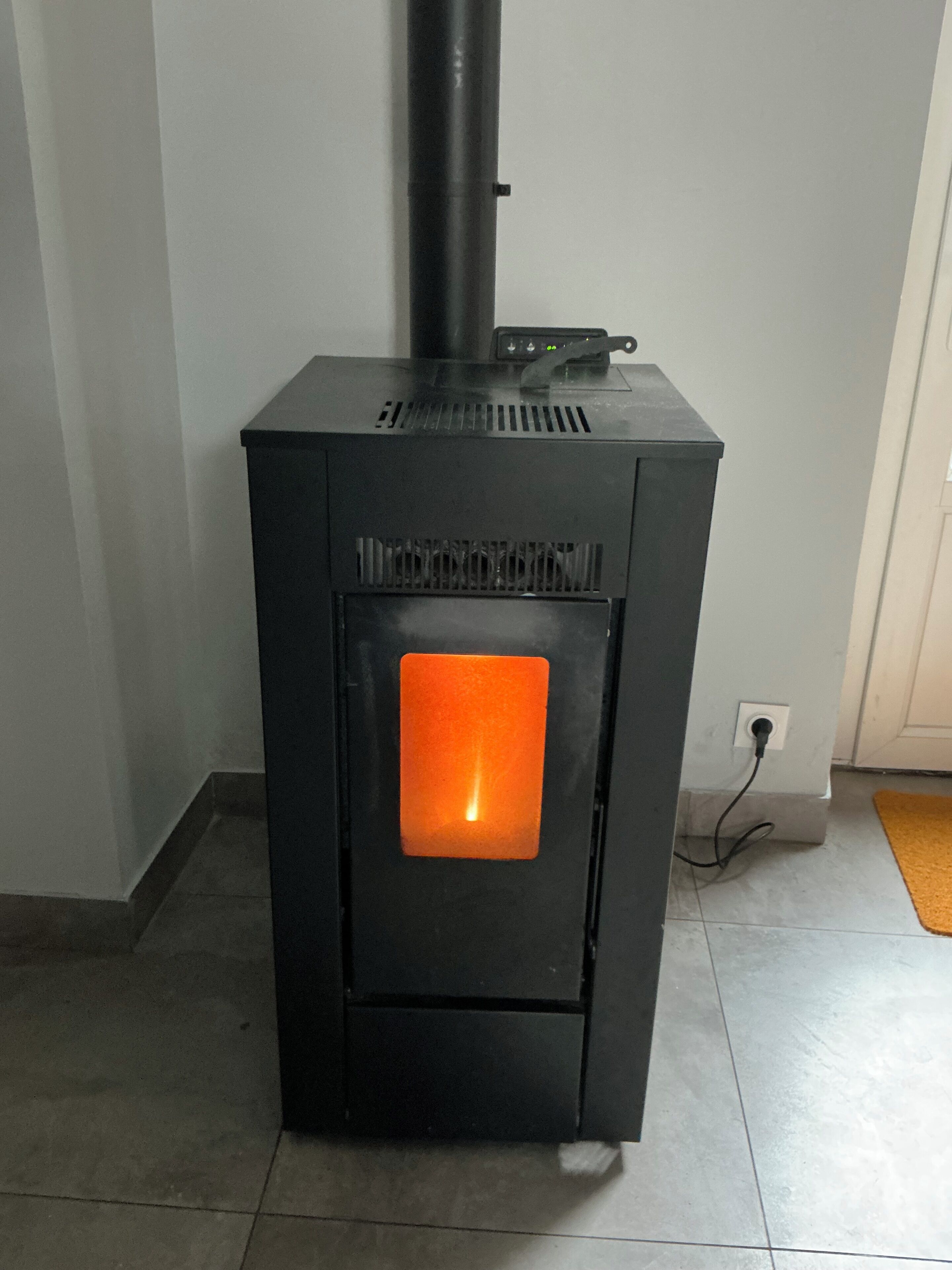 Una stufa a legna in legno nero con sportello in vetro arancione, un comignolo nero e una presa di corrente sulla parete.