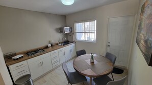 Une cuisine compacte équipée d’une cuisinière à gaz, d’un four à micro-ondes et d’un évier. On y trouve une table à manger avec quatre chaises et une plante en pot. Une poubelle est visible, et la pièce est dotée d’une porte blanche et d’une fenêtre avec des stores.