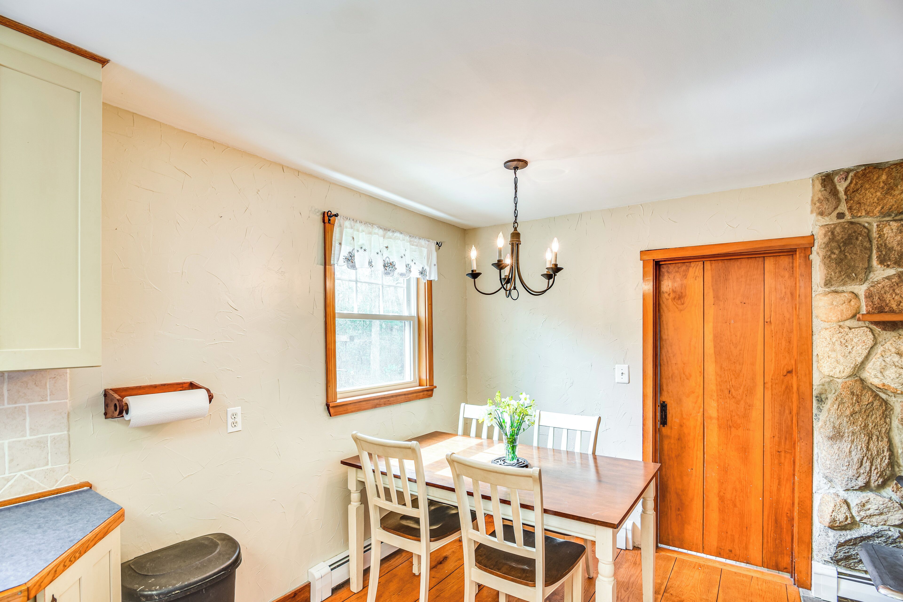 A dining area with a wooden table, white chairs, a chandelier, a window with a floral curtain, and a stone wall.