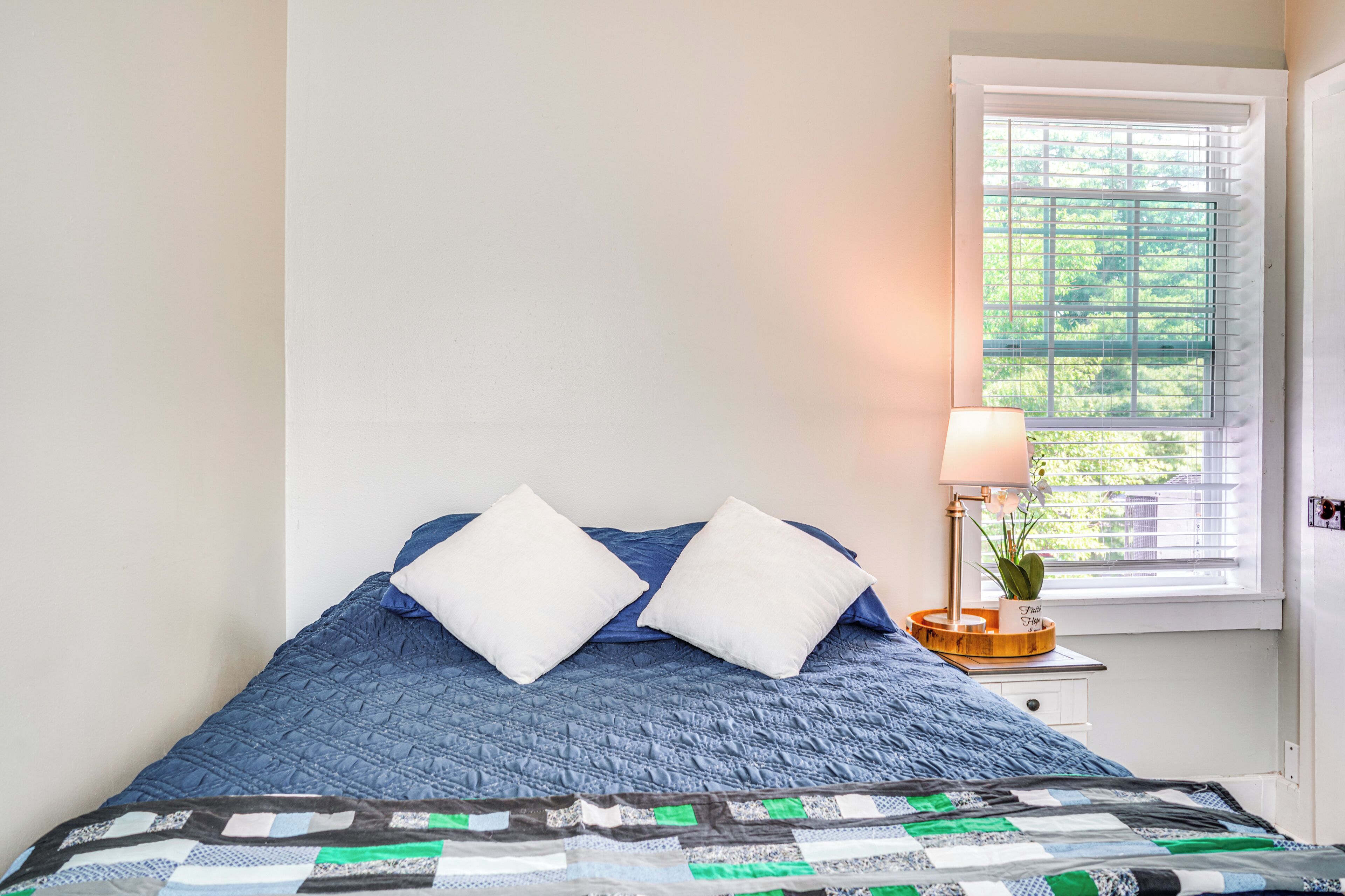A bed with a blue quilt, two white pillows, a bedside table with a lamp, and a window with blinds.