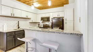 Una cocina moderna con armarios blancos, encimeras de granito, un refrigerador de acero inoxidable y un microondas. Hay dos lámparas colgantes y un taburete de bar.