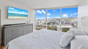 A bedroom with a large bed, a dresser, and a city view through a large window. - Beach 180 degrees City and Mountain  And centrally located Kitchen fireworks (Honolulu)