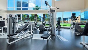 A gym with various exercise machines, large windows, and a view of palm trees and buildings. - Beach 180 degrees City and Mountain  And centrally located Kitchen fireworks (Honolulu)
