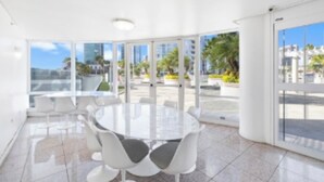 A modern indoor space with a round glass table and white chairs, large windows offering a view of palm trees and buildings. - Beach 180 degrees City and Mountain  And centrally located Kitchen fireworks (Honolulu)