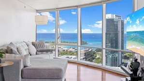 A modern living room with a large window overlooking a beach and ocean, a grey sofa, and a lamp. - Beach 180 degrees City and Mountain  And centrally located Kitchen fireworks (Honolulu)