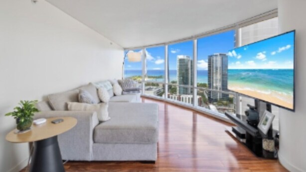 A modern living room with a large window offering a city view, a comfortable sofa, and a flat-screen TV. - Beach 180 degrees City and Mountain  And centrally located Kitchen fireworks (Honolulu)