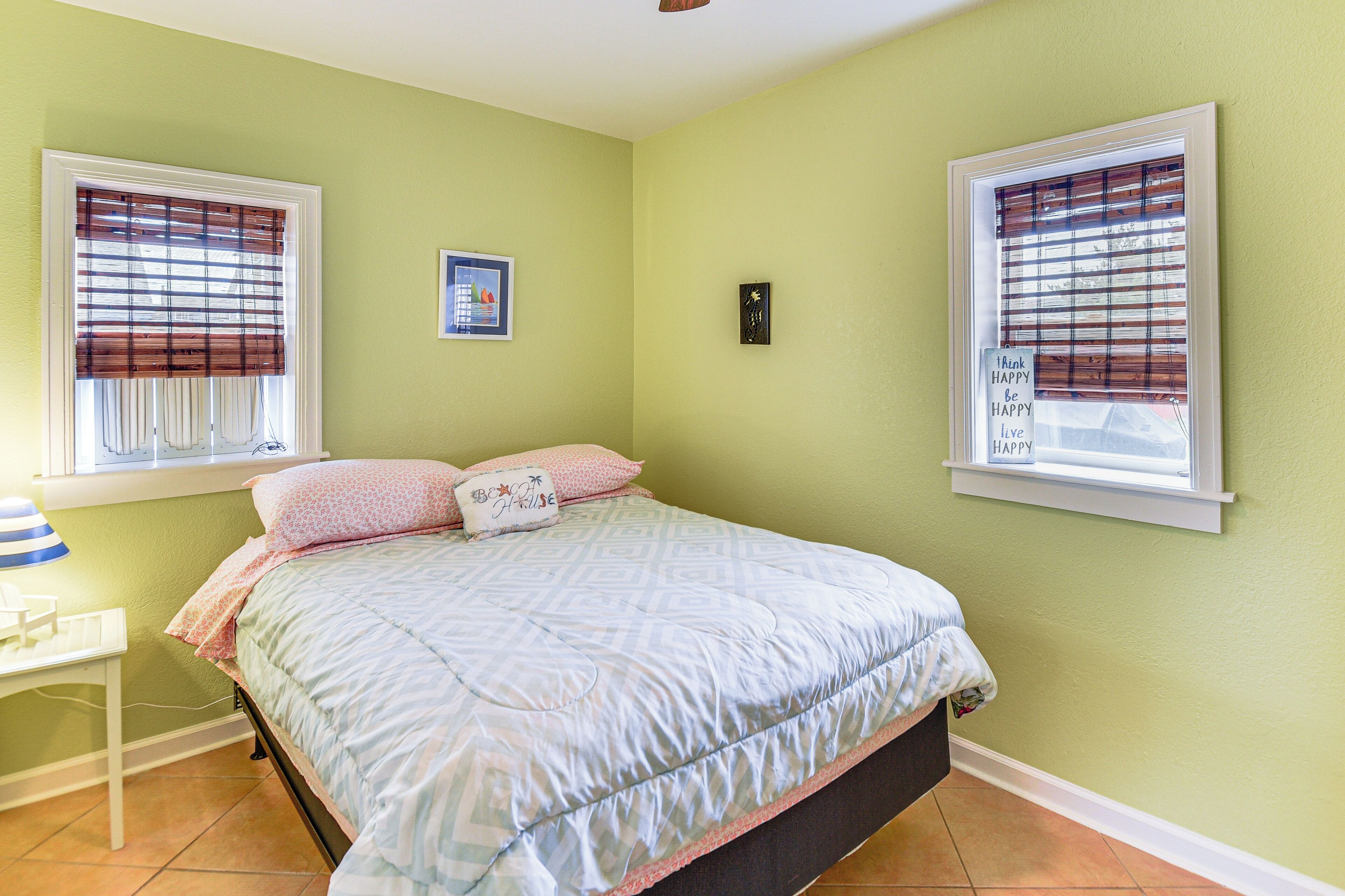 A bedroom with a bed, two windows with blinds, a bedside table, and a wall clock.