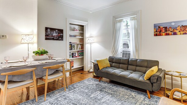 Ein Wohnzimmer mit grauem Sofa, Holzstühlen, einem Esstisch für vier Personen, einem Bücherregal und einem Stadtbild.
