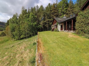 A rustic cabin with a stone exterior, surrounded by dense forest and a well-maintained lawn. - Creag Bhalg (Ballater)