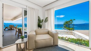 Una sala moderna con un sofá beige, una mesita redonda con una planta y vista al océano a través de grandes ventanales.
