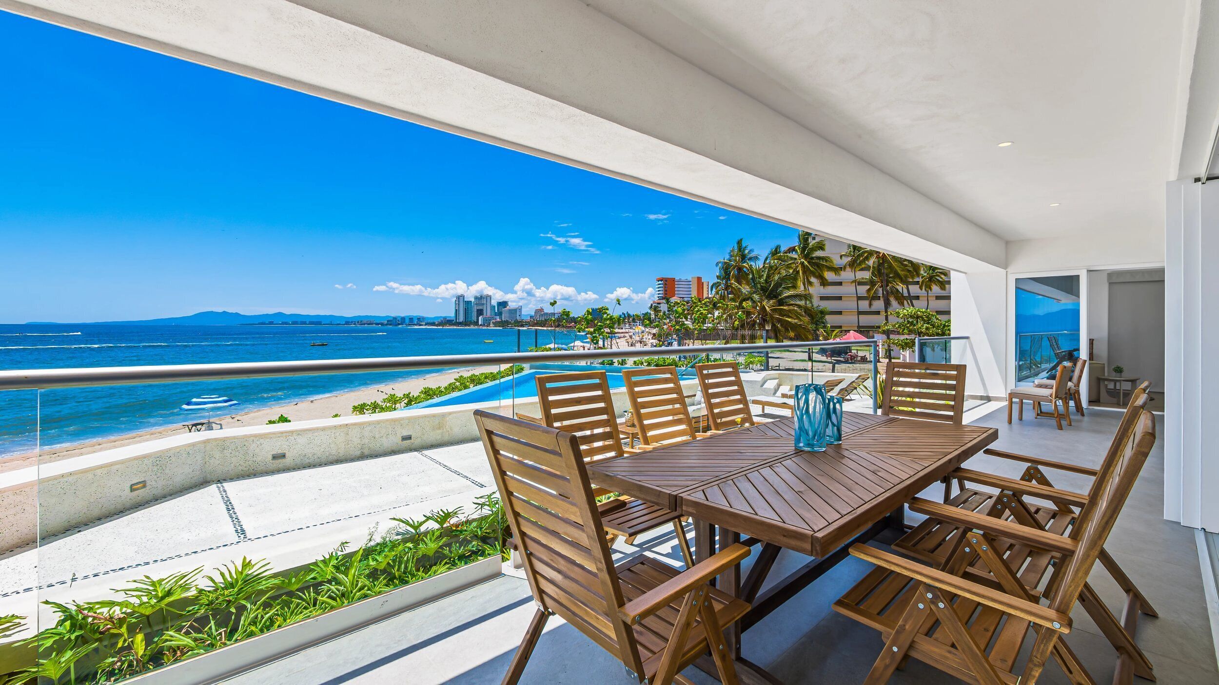 Une table en bois, entourée de chaises, sur un balcon donnant sur une plage et l’océan.